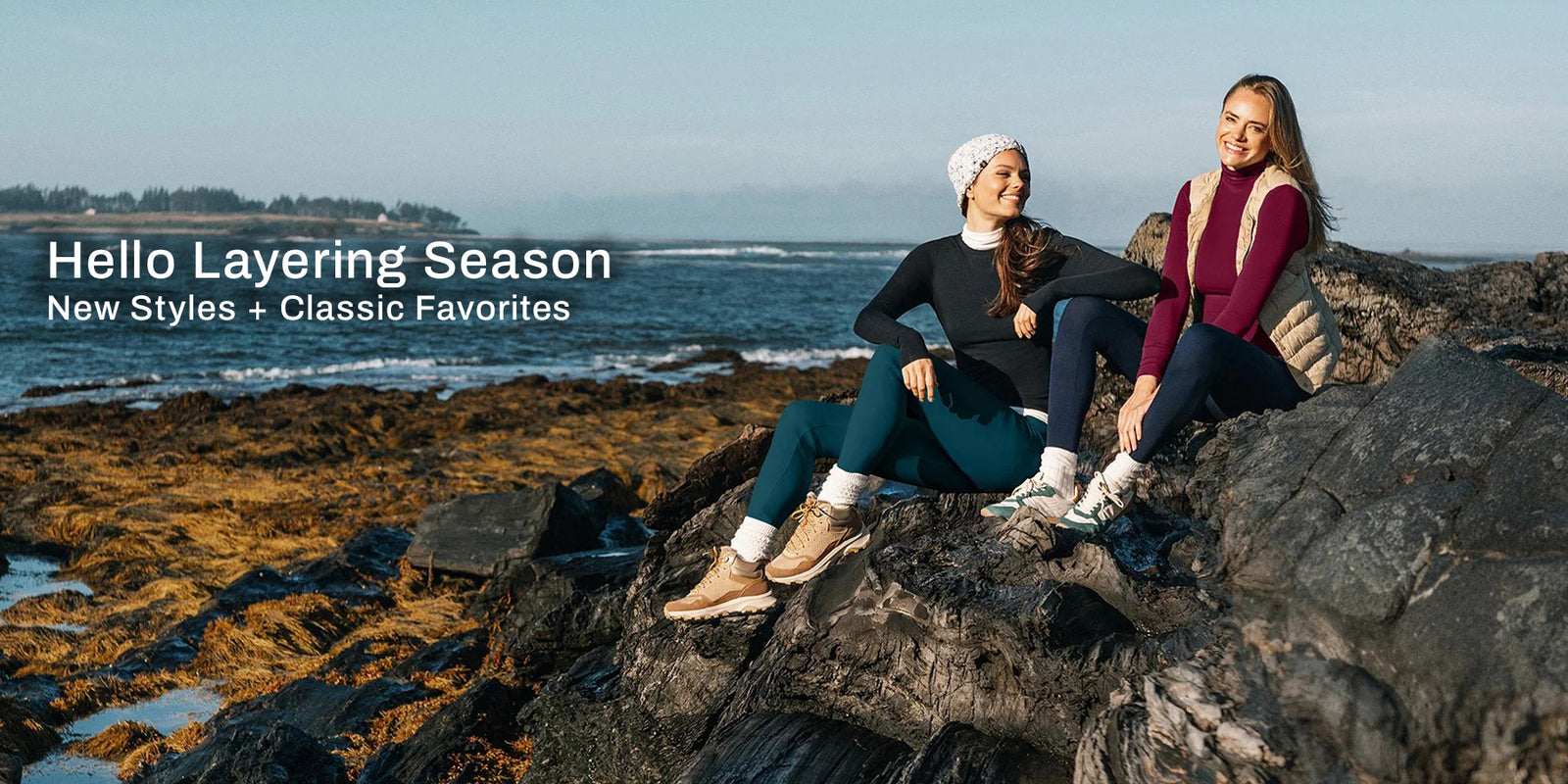 Two women sitting on dark rocks by the ocean, wearing layered winter outfits. The left woman wears a black top, teal leggings, white socks, and tan boots, while the right woman wears a burgundy top, dark leggings, and a beige vest.