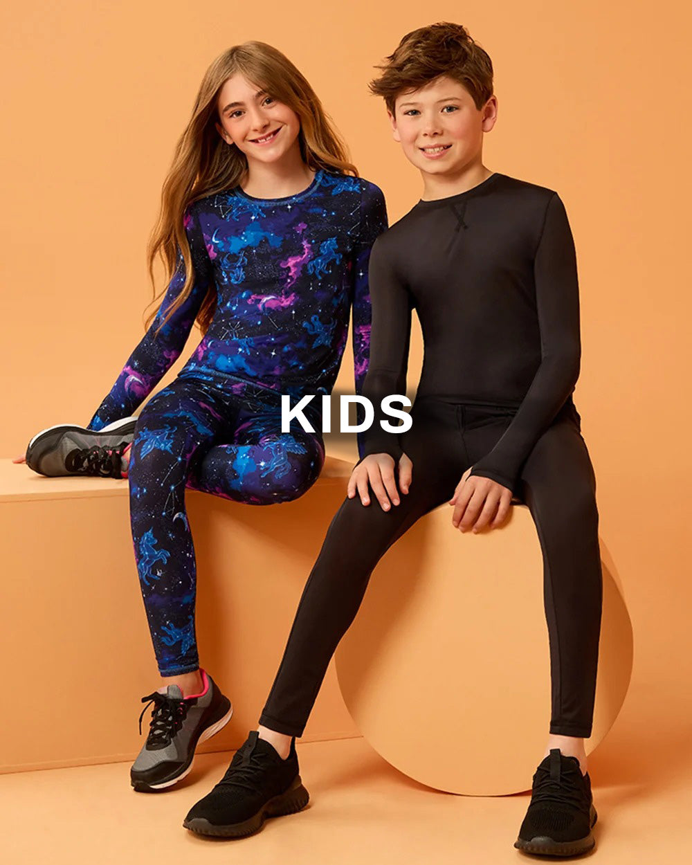 Two children modeling matching long-sleeve athletic sets in a studio setting. The girl wears a blue and purple galaxy print one-piece with matching leggings, and the boy wears a solid black one-piece with matching leggings.