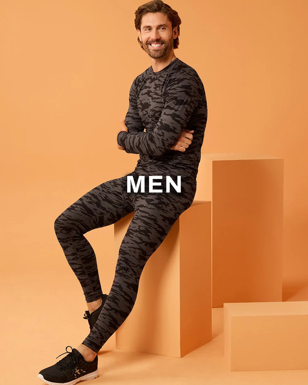 A man wearing a dark gray and black camouflage long-sleeve athletic top and matching leggings, posing on orange display blocks against a solid orange background. The word 'MEN' is overlaid in white text across the center.